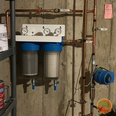 A home water filtration system installed on a basement wall, featuring two large blue filter housings with pressure gauges mounted above each. Copper plumbing lines run vertically and horizontally, connecting to a water pump on the right. The setup is mounted on a concrete wall near a shelving unit with paint cans and other supplies. A circular logo is visible in the bottom right corner.