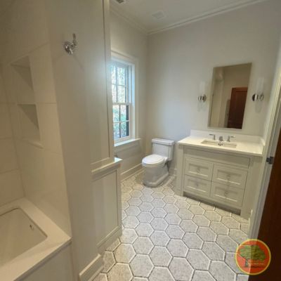A bright, modern bathroom with a clean, neutral color palette. The floor features large hexagonal tiles with dark grout lines. On the right side, there is a white vanity with double drawers, a rectangular sink, a wall-mounted mirror, and sconces on either side. A toilet is positioned between the vanity and a large window. To the left is a built-in bathtub with tile shelving and a chrome shower fixture. Recessed lighting is installed in the ceiling.